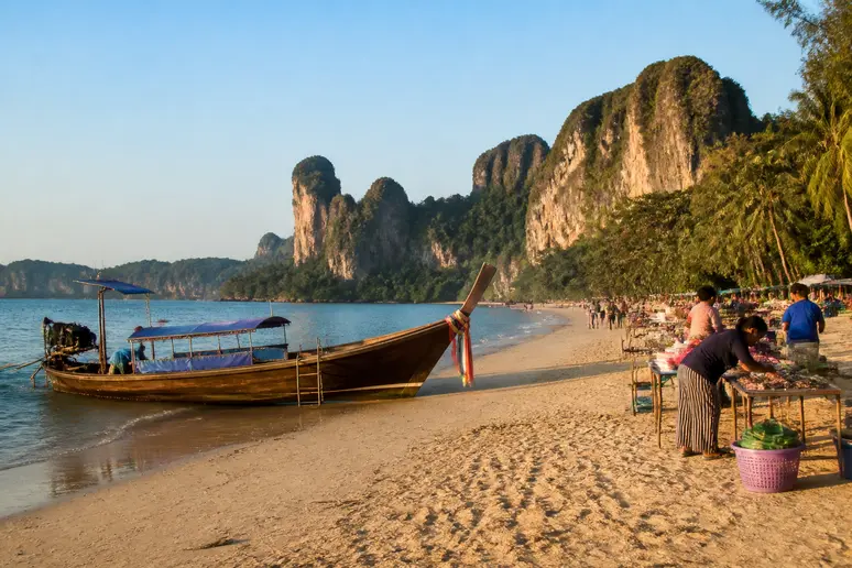 Ein Longtail-Boot am Ao Nang Beach mit dramatischen Kalksteinfelsen im Hintergrund, Lokale bereiten sich auf den Abend vor.