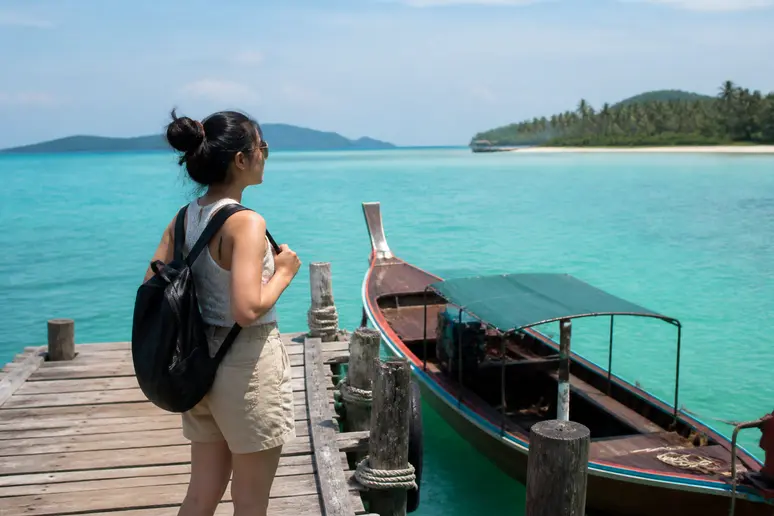 Eine junge thailändische Frau steht an einem kleinen Pier auf Koh Kood und blickt auf das türkisfarbene Meer, bereit für die Weiterreise.
