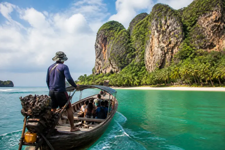 Ein lokaler Fischer steuert ein traditionelles Longtail-Boot durch das klare Wasser auf dem Weg nach Rai Leh Beach in Thailand, umgeben von beeindruckenden Kalksteinfelsen.