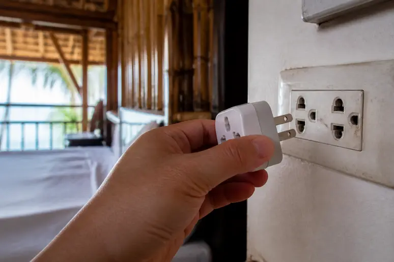 Ein Reiseadapter mit flachen Pins wird in eine Steckdose in einem thailändischen Guesthouse gesteckt, schützt vor Abnutzung.