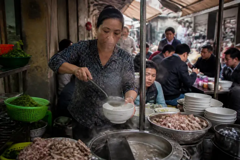 Eine Garküche in Hanoi, Vietnam, mit einer Frau, die dampfende Pho-Schüsseln an Einheimische ausgibt, authentisches kulinarisches Erlebnis.