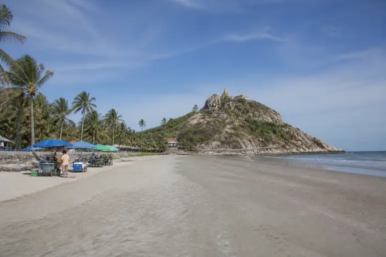 Weitläufiger Khao Takiab Beach in Hua Hin mit Felsen, Tempel und lokalen Verkäufern im Hintergrund.