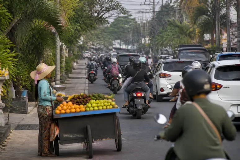 Verkehr in Seminyak Bali: Motorroller und Autos auf belebter Straße, Straßenhändler mit Obst.