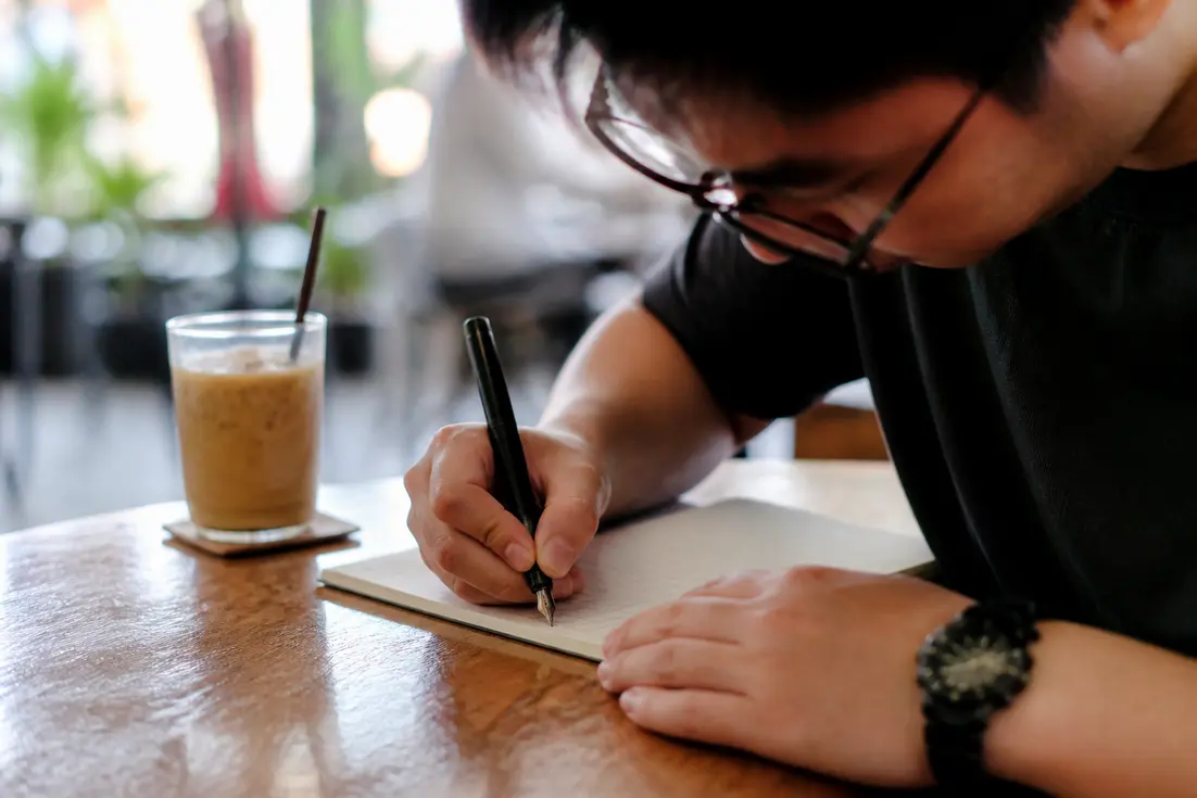 Eine Person sitzt an einem Holztisch und übt das Thailändische Schrift lesen und schreiben lernen mit einem Notizbuch.