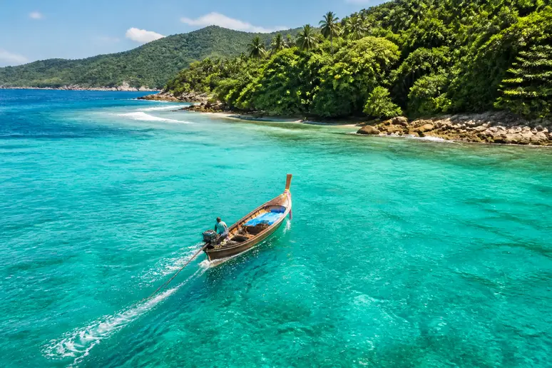Ein Longtailboot nähert sich dem The Treasure Koh Madsum Resort, umgeben von türkisfarbenem Wasser und tropischer Inselvegetation, die The Treasure Koh Madsum Erfahrungen widerspiegelt.