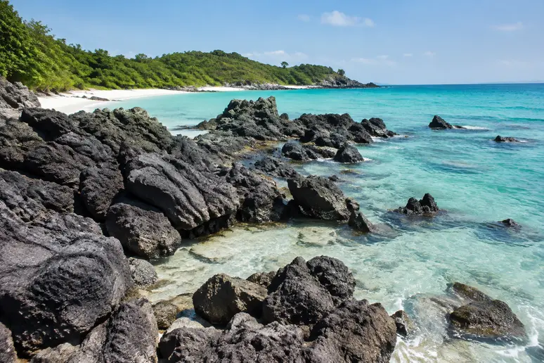 Dunkle Felsblöcke und karge Küstenlinie an der Küste von Koh Kham island bei klarem Wasser.